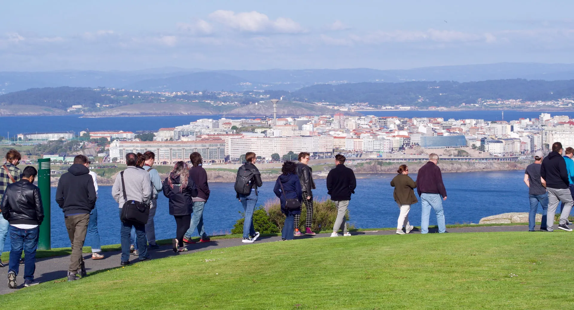 A photo showing a group of people walking from left to right on a hill overlooking a city on a peninsula, with blue water, rolling green hills and slightly cloudy sky.